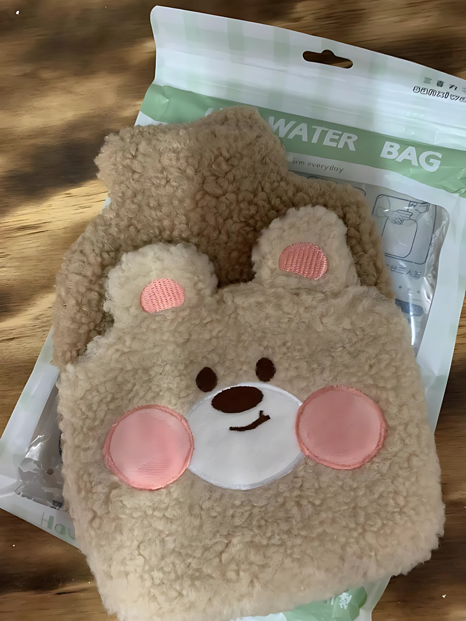Bolsa térmica de felpa beige con diseño de oso, orejas y mejillas rosadas, sobre su empaque que indica "WATER BAG".