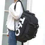 Bolso deportivo negro con diseño T60 en blanco, usado como mochila por persona con sudadera blanca y jeans. Incluye compartimento lateral para calzado.