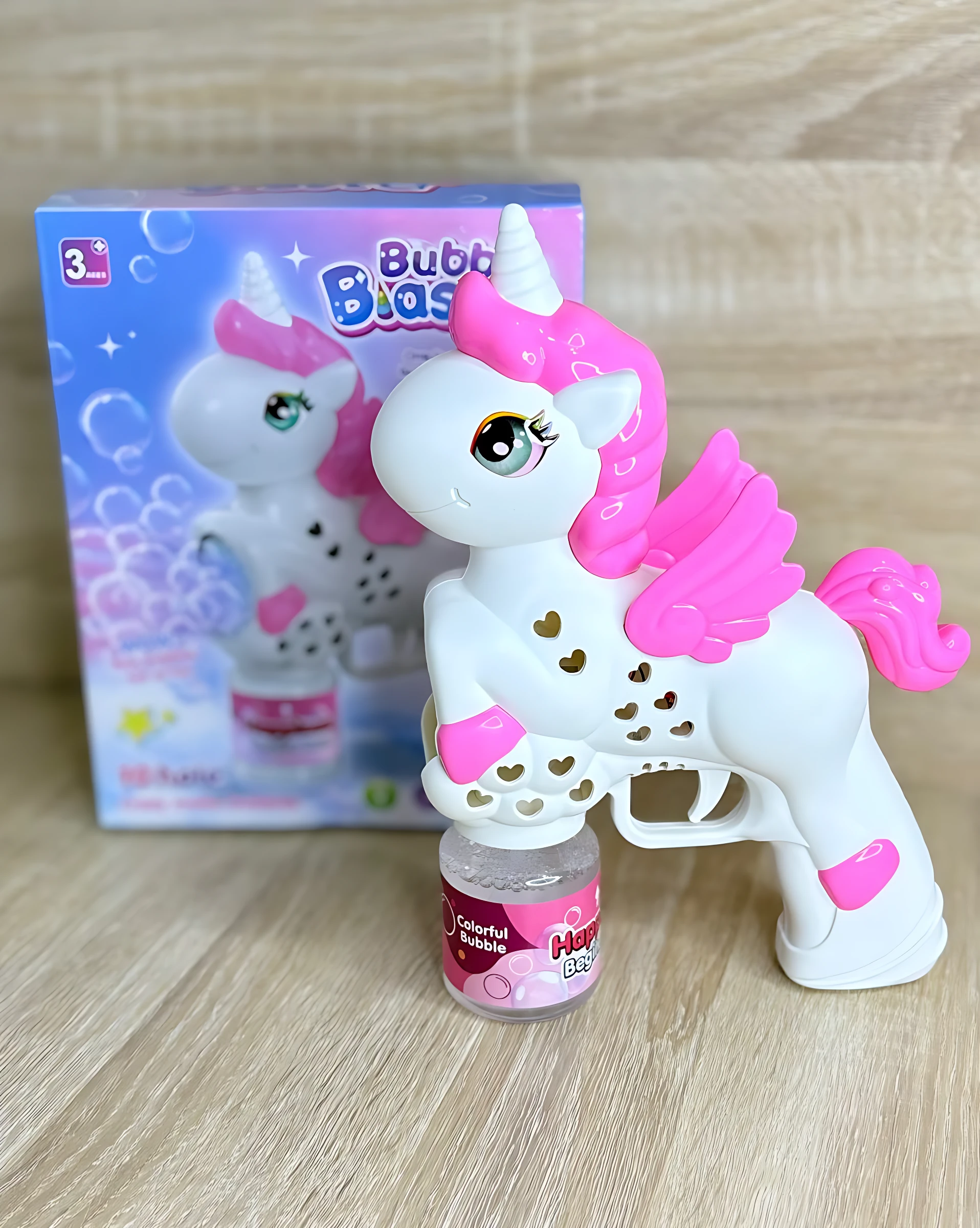 Burbujero automático en forma de unicornio blanco con detalles rosas, alas y cuerno, junto a su caja y frasco de líquido.