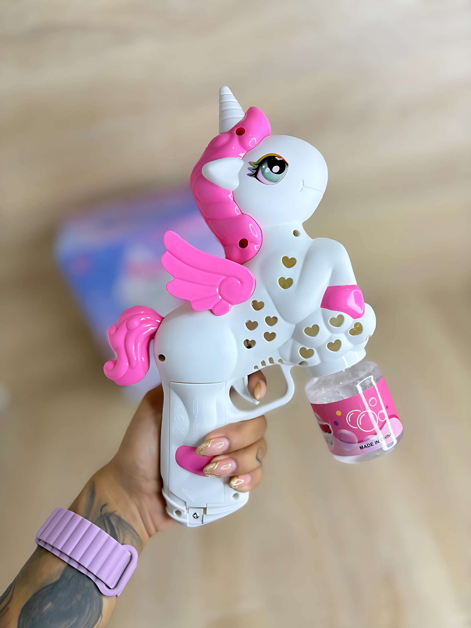 Burbujero automático en forma de unicornio blanco con detalles rosas y alas, accionado por mano, con depósito de líquido transparente y tapa rosa.