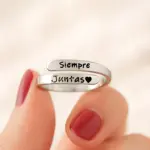Anillo ajustable de plata con dos bandas grabadas que dicen Siempre Juntas y un corazón, sostenido entre los dedos con uñas pintadas de rojo.