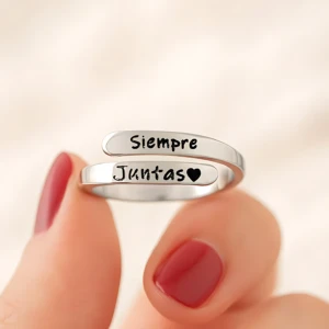 Anillo ajustable de plata con dos bandas grabadas que dicen Siempre Juntas y un corazón, sostenido entre los dedos con uñas pintadas de rojo.