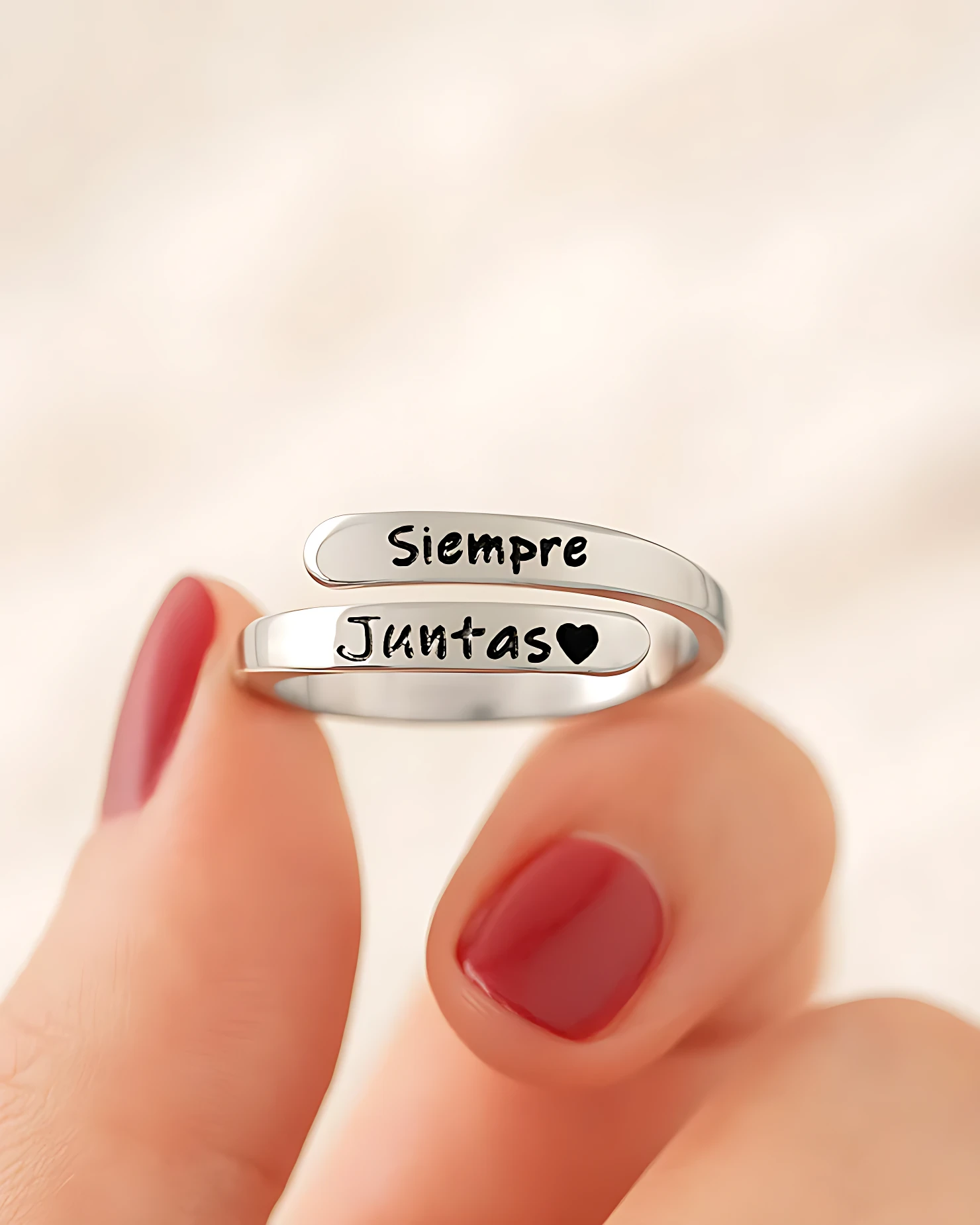 Anillo ajustable de plata con dos bandas grabadas que dicen Siempre Juntas y un corazón, sostenido entre los dedos con uñas pintadas de rojo.