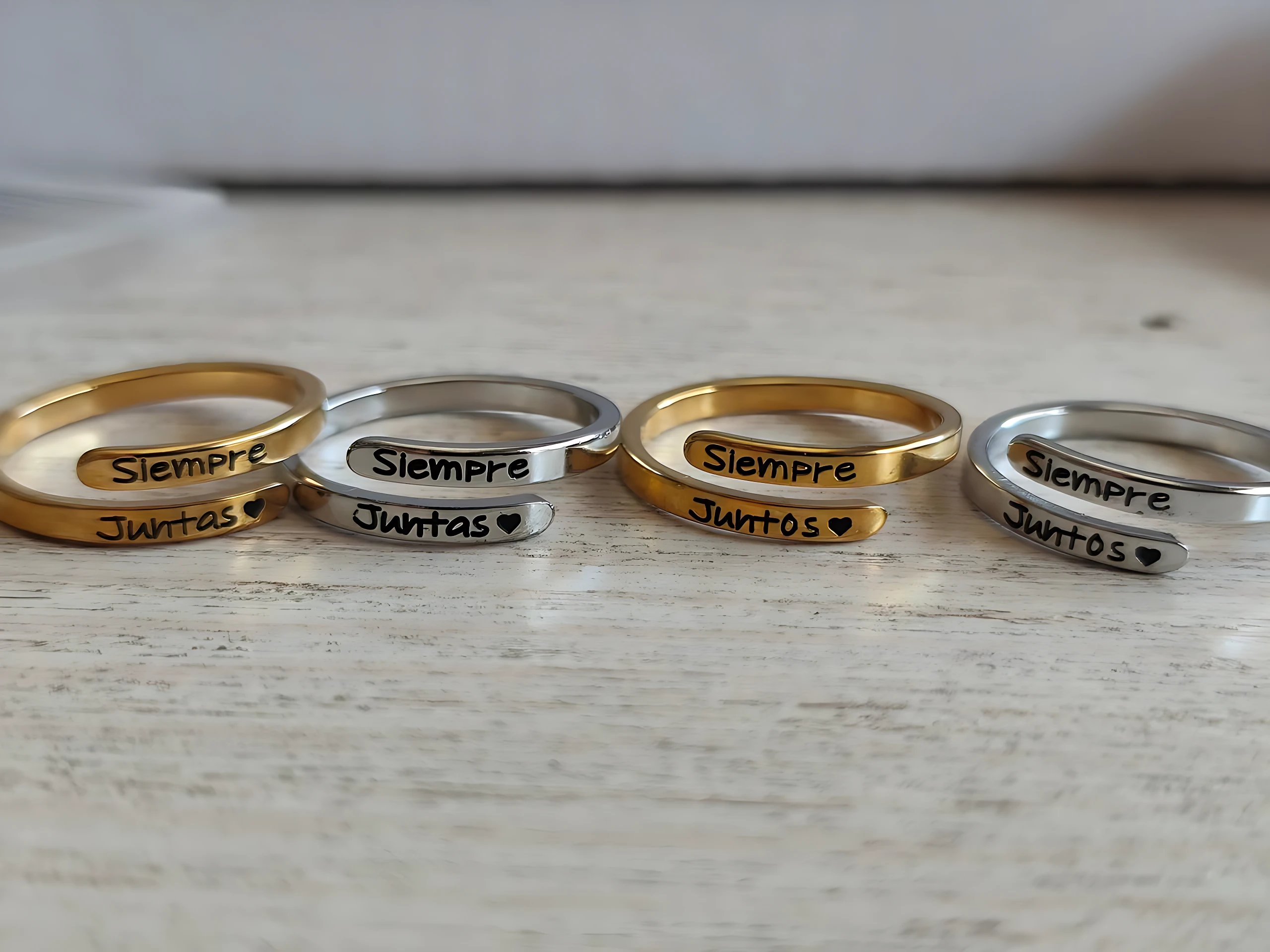 Cuatro anillos ajustables de oro y plata con la inscripción Siempre Juntas y corazón grabado, dispuestos en fila sobre superficie clara.