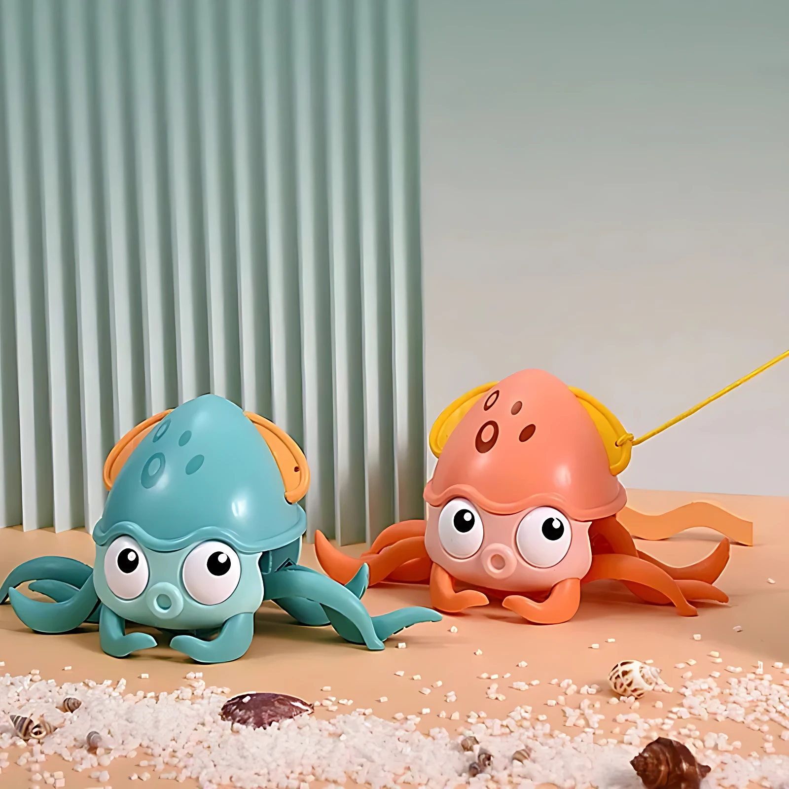 Dos juguetes de calamar caminante interactivos, uno azul y otro naranja, con ojos grandes y tentáculos flexibles, sobre arena con conchas pequeñas.