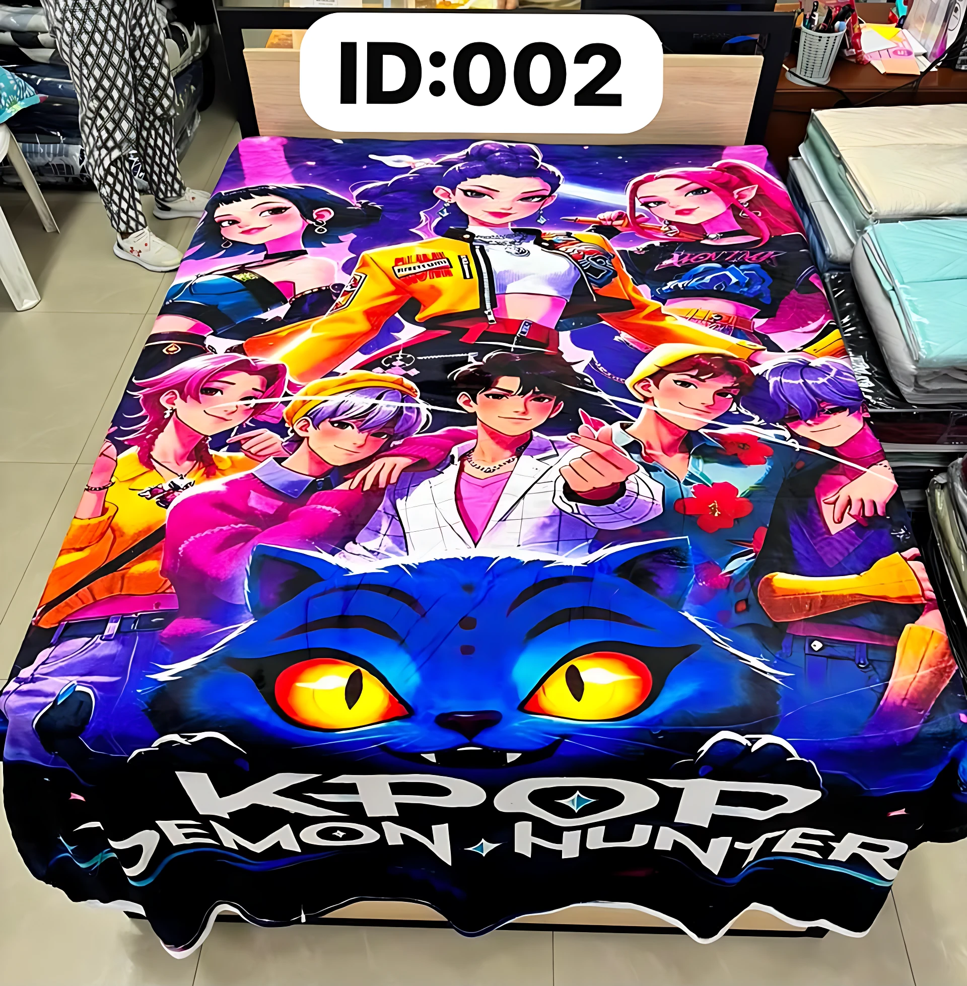 Cobija digitalizada con estampado de personajes KPOP y gato azul brillante, sobre cama en tienda.