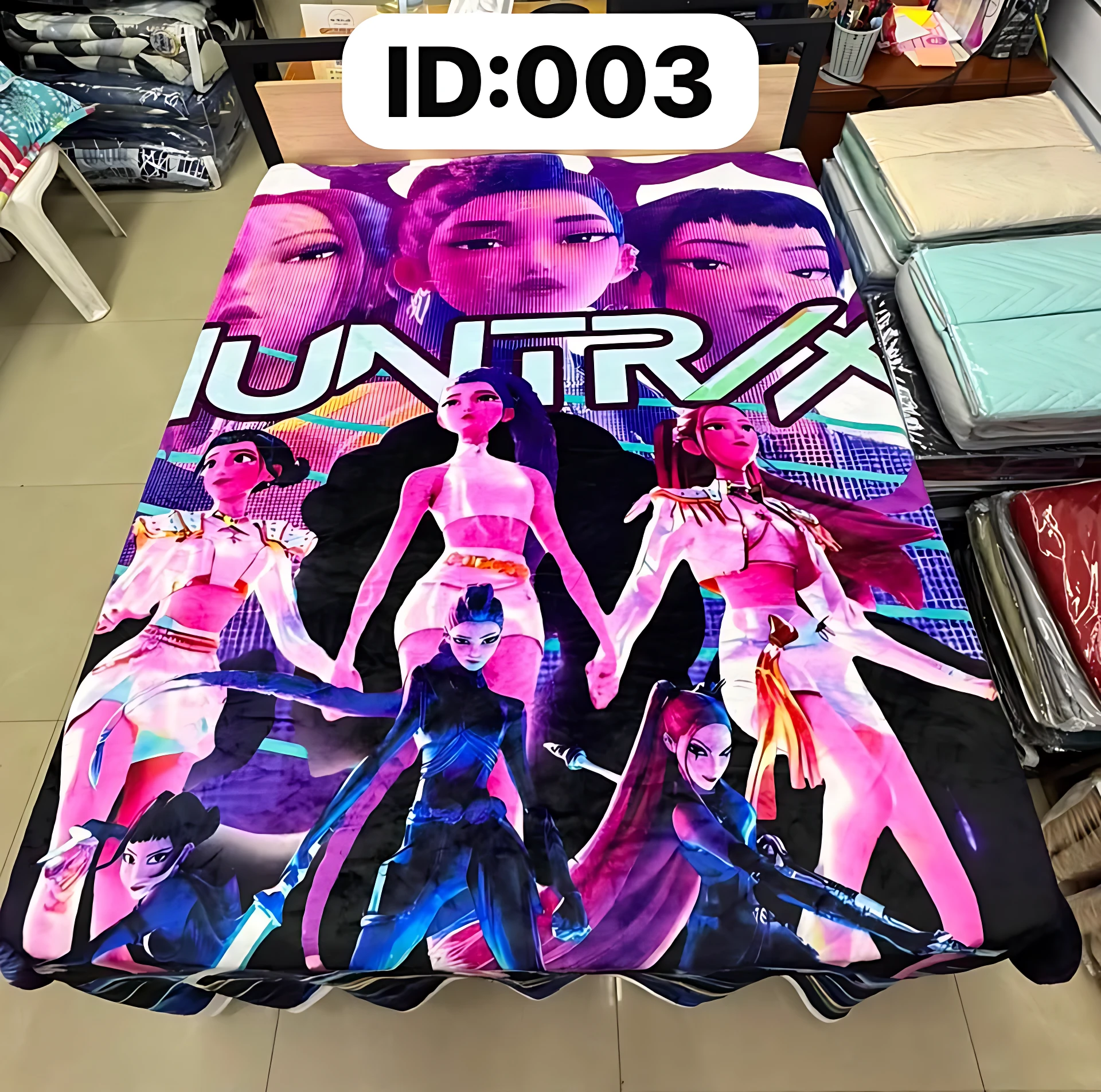 Cobija digitalizada con estampado de personajes animados en tonos neón sobre fondo negro, extendida sobre cama en ambiente de tienda.