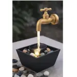 Grifo dorado clásico con agua cayendo sobre cuenco negro con luces LED, rodeado de piedras decorativas en fuente cuadrada, ideal para ambiente relajante.