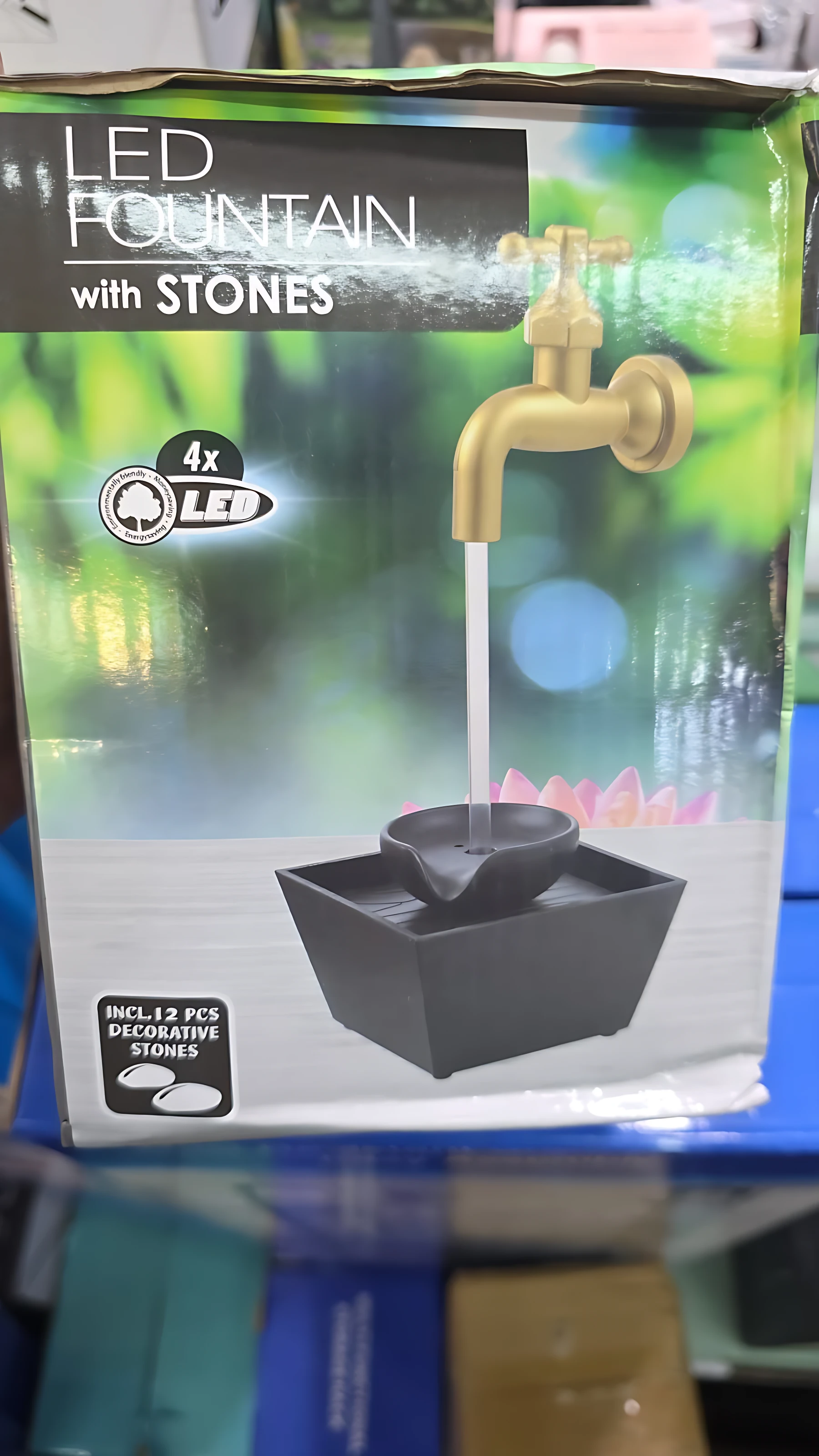 Fuente decorativa LED con grifo dorado y piedras negras, en caja con fondo verde borroso. Incluye luz nocturna y ambiente relajante.