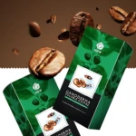 Dos paquetes verdes de café soluble con extracto de ganoderma, rodeados de granos de café y hojas, sobre fondo marrón.