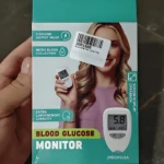 Caja de glucometro digital con pantalla grande, mostrando dispositivo blanco y lector de gota de sangre en mano de mujer sonriente.