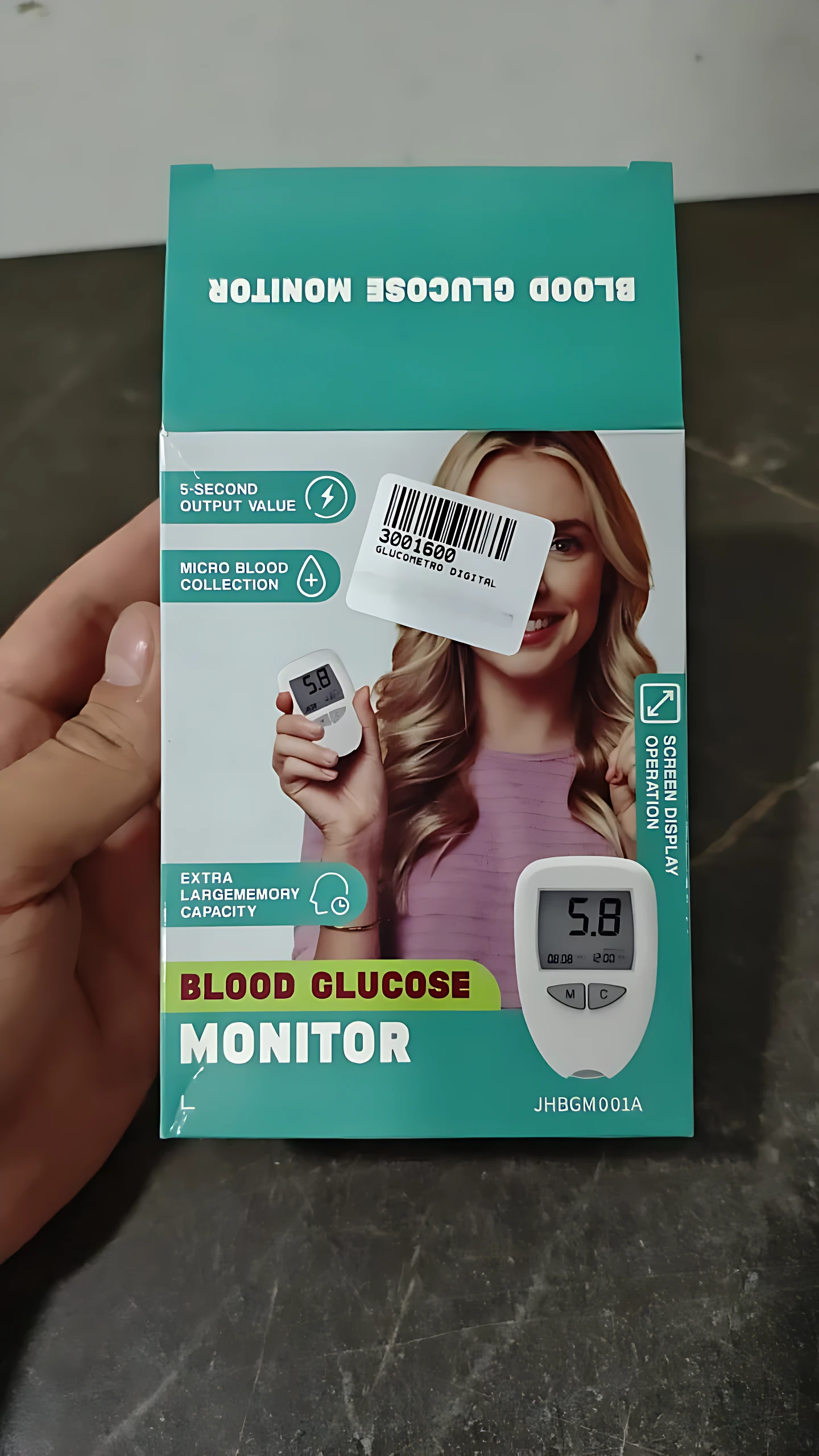 Caja de glucometro digital con pantalla grande, mostrando dispositivo blanco y lector de gota de sangre en mano de mujer sonriente.