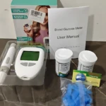 Kit de glucómetro digital con pantalla grande, caja, manual, dos frascos de tiras, estuche con sensor y bolsa de lancetas azules sobre superficie oscura.