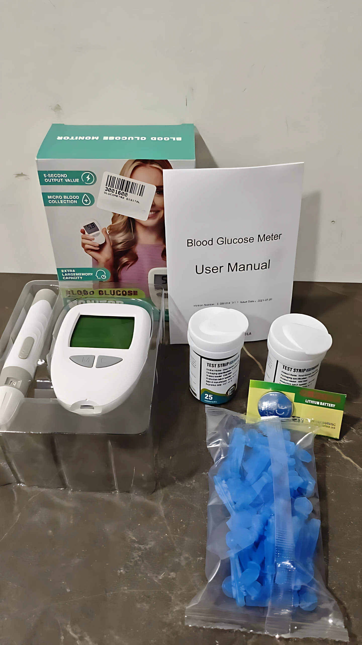 Kit de glucómetro digital con pantalla grande, caja, manual, dos frascos de tiras, estuche con sensor y bolsa de lancetas azules sobre superficie oscura.