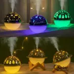 Humificador lámpara planeta 3D con proyección de luz: muestra tres unidades en colores amarillo, morado y verde, con patrones estelares y navideños; abajo, tres más sobre soportes de madera, uno con luces de hadas, todos emitiendo vapor.