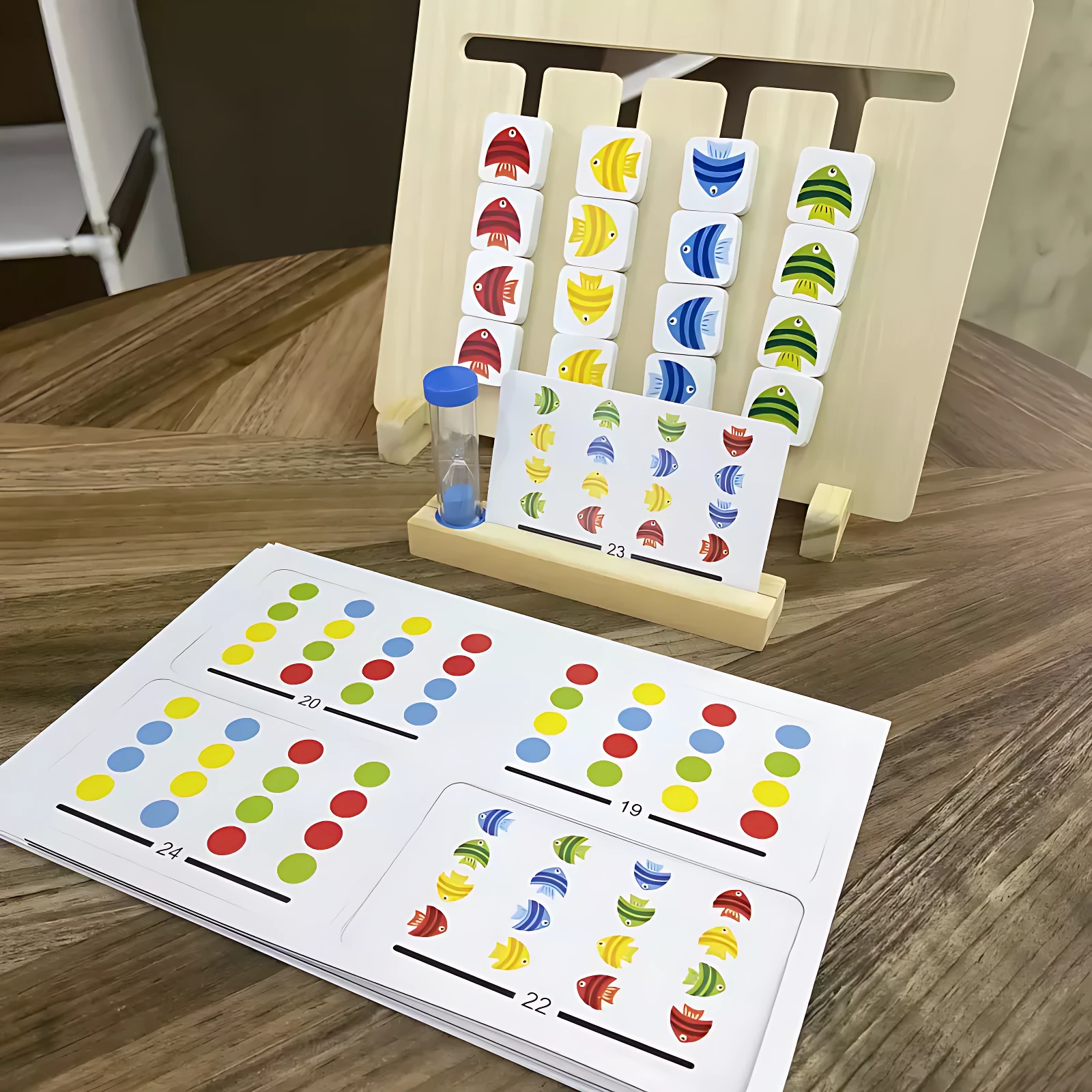 Juego didáctico de madera Montessori con fichas de peces multicolores, tarjetas numeradas y temporizador azul sobre mesa de madera.