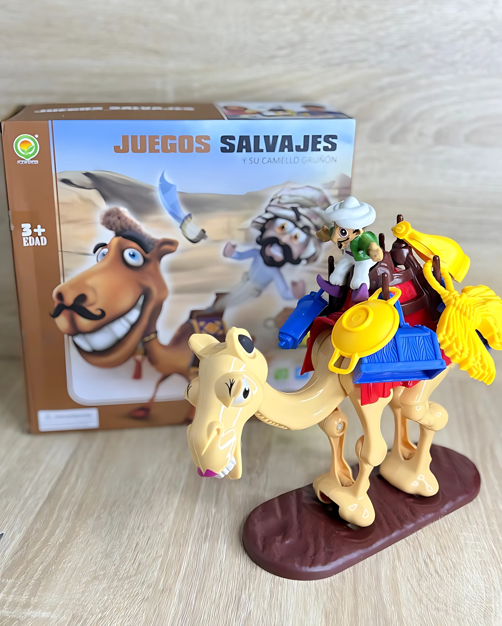 Juego de mesa familiar con figura de camello beige sobre base marrón, acompañado de personajes y accesorios coloridos, junto a su caja original.