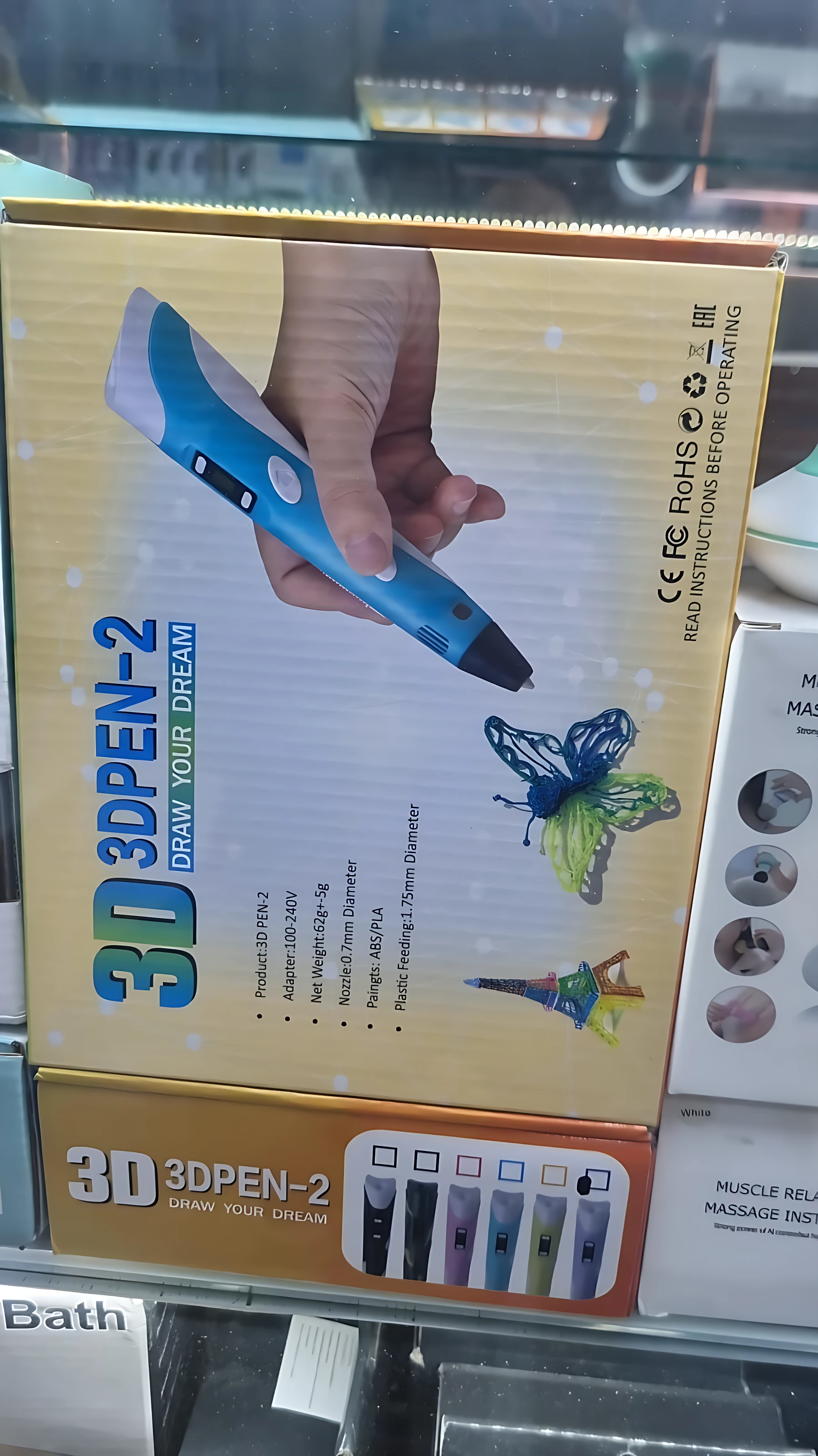 Lápiz de impresión 3D portátil en su caja amarilla, mostrando mano sosteniendo el dispositivo azul con punta negra, junto a ilustraciones de mariposa y torre Eiffel. Incluye accesorios visibles en la parte inferior.