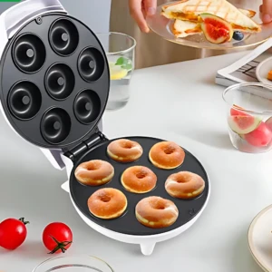 Máquina para hacer donas con siete moldes negros, abierta sobre mesa blanca, mostrando donas horneadas y accesorios de cocina al fondo.