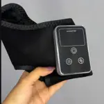 Masajeador inalámbrico para tobillo en color negro, con panel de control digital y botones de encendido, calor y vibración, sostenido en la mano sobre fondo gris.