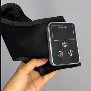 Masajeador inalámbrico para tobillo en color negro, con panel de control digital y botones de encendido, calor y vibración, sostenido en la mano sobre fondo gris.