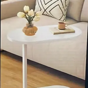 Mesa auxiliar blanca con base ovalada y superficie redondeada, sobre sofá beige con cojín geométrico; incluye jarrón con tulipanes y taza sobre libro.