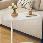 Mesa auxiliar blanca con base en forma de U, superficie redondeada y acabado liso, sobre piso de madera con sofá beige al fondo.