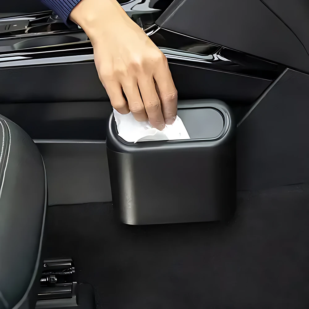 Basurero negro para carro, con tapa abatible y mano retirando papel higiénico, instalado en piso de vehículo.