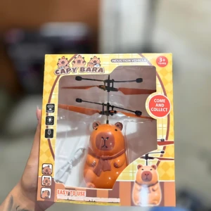 Muñeco volador capibara naranja en empaque, con hélice visible y etiqueta de 3+ años.