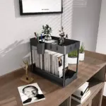 Organizador de dos niveles en color negro, con estructura cuadrada y rejilla metálica, exhibido sobre una mesa de madera con libros, plantas y accesorios decorativos.