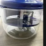 Picadora manual multifuncional con base transparente y tapa azul, vista frontal mostrando cuchillas de acero inoxidable en posición de corte.