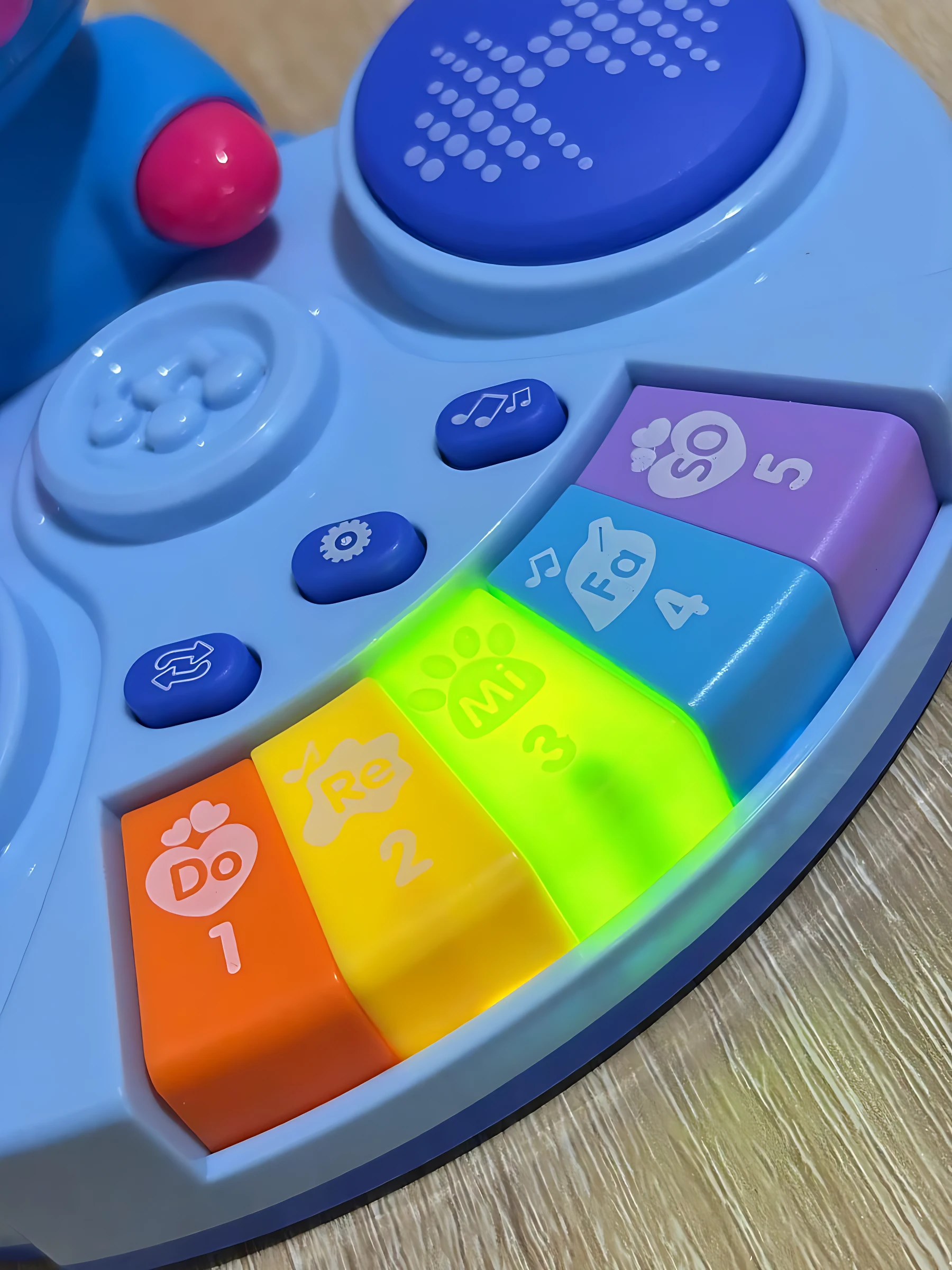 Piano educativo para niños con teclas de colores Do a Sol, botones de función y panel de control azul, visto en primer plano sobre superficie de madera.