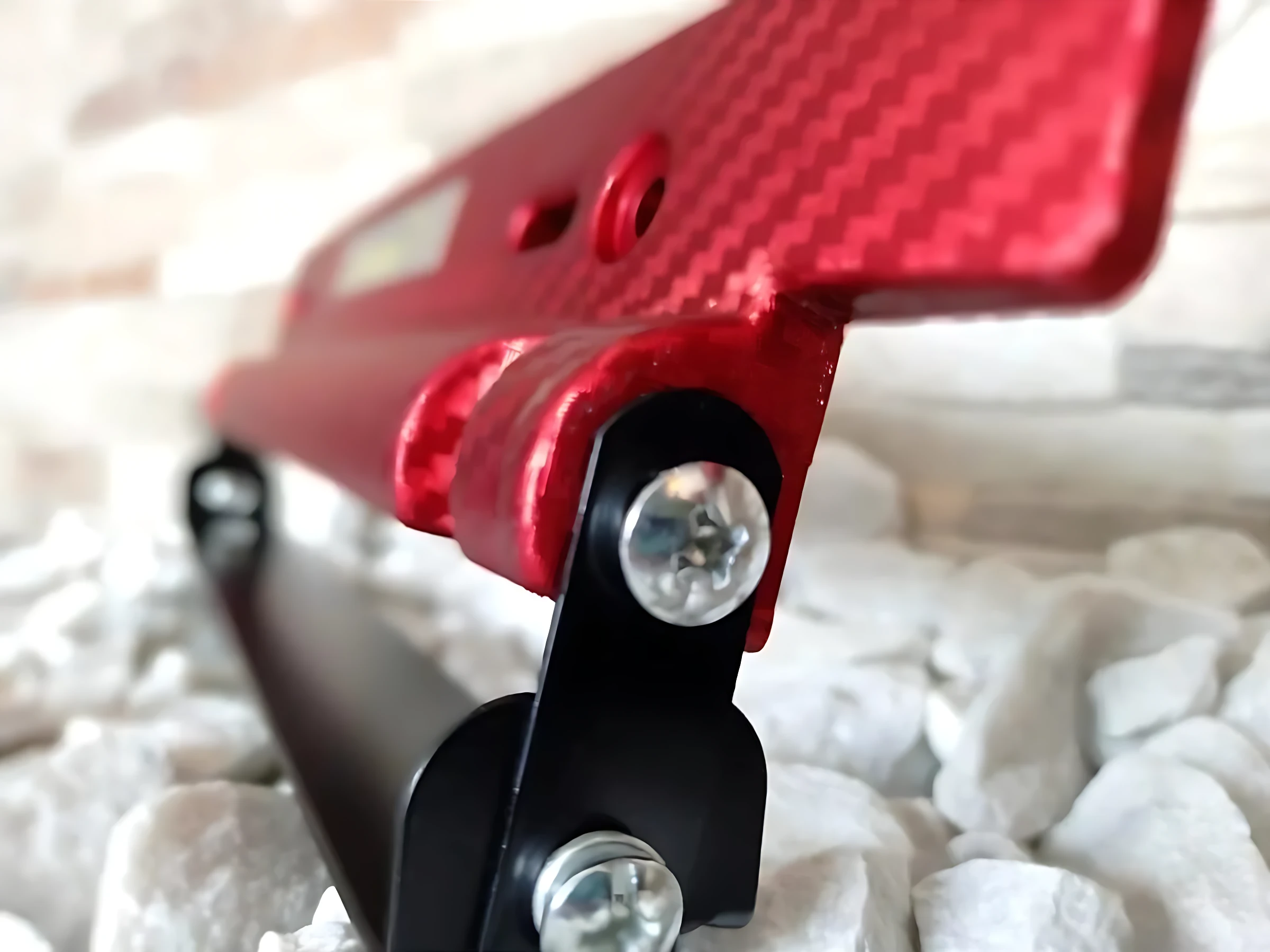 Porta placa fibra carbono rojo con platina metálica, detalle de montaje con tornillos visibles sobre base negra, textura de fibra visible y acabado brillante.