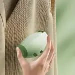 Mano sosteniendo quitamotas ergonómico en color verde claro, deslizándose sobre tela beige con textura visible, eliminando pelusas de prendas delicadas.