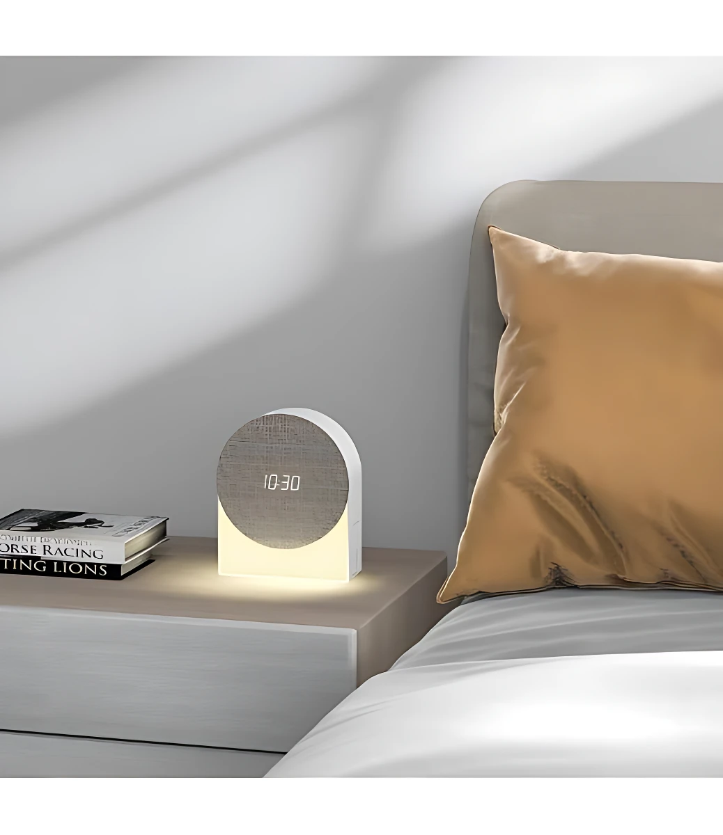 Reloj despertador inteligente con luz nocturna y sonidos de naturaleza, mostrando hora 10:30 sobre mesita de noche junto a cama con almohada beige.