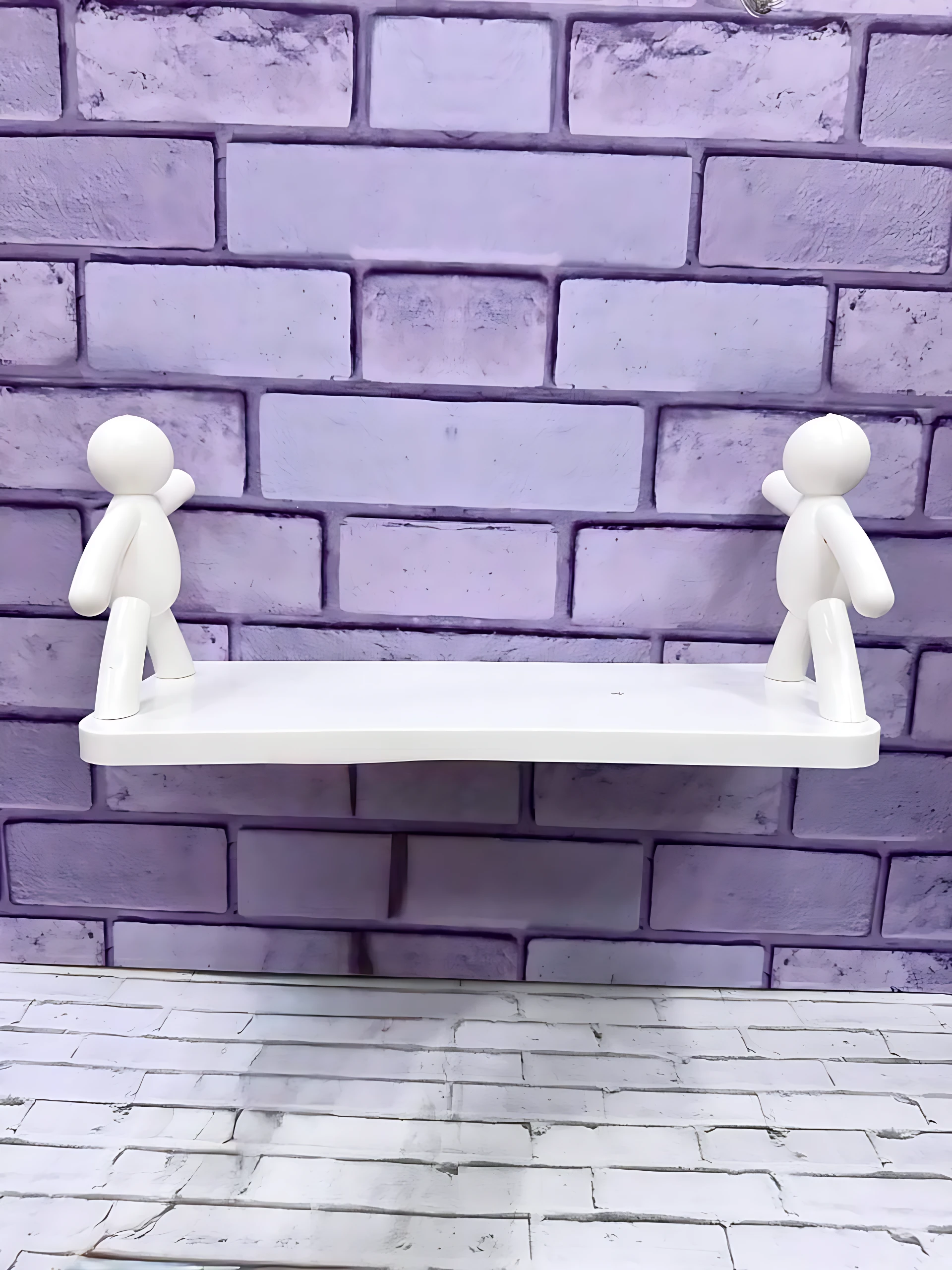 Repisa blanca con dos figuras humanas de estilo minimalista, montada sobre pared de ladrillo pintado en tono lavanda. Ideal para exhibir objetos pequeños en espacios reducidos.