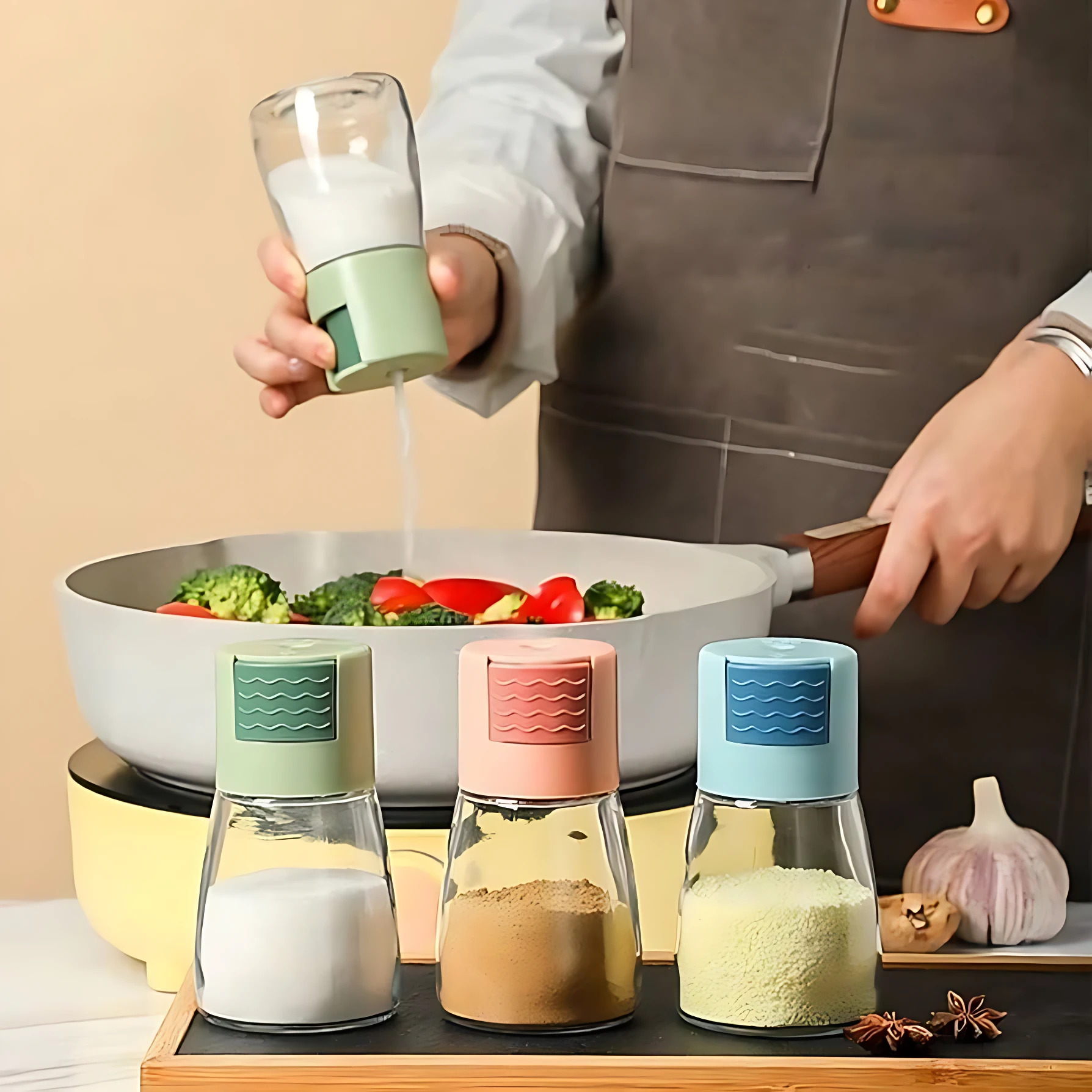 Set de tres saleros de vidrio con tapa de plástico en verde, rosa y azul, cada uno con medidor de gramos visible, sobre tabla de madera junto a sartén con vegetales y ajo.