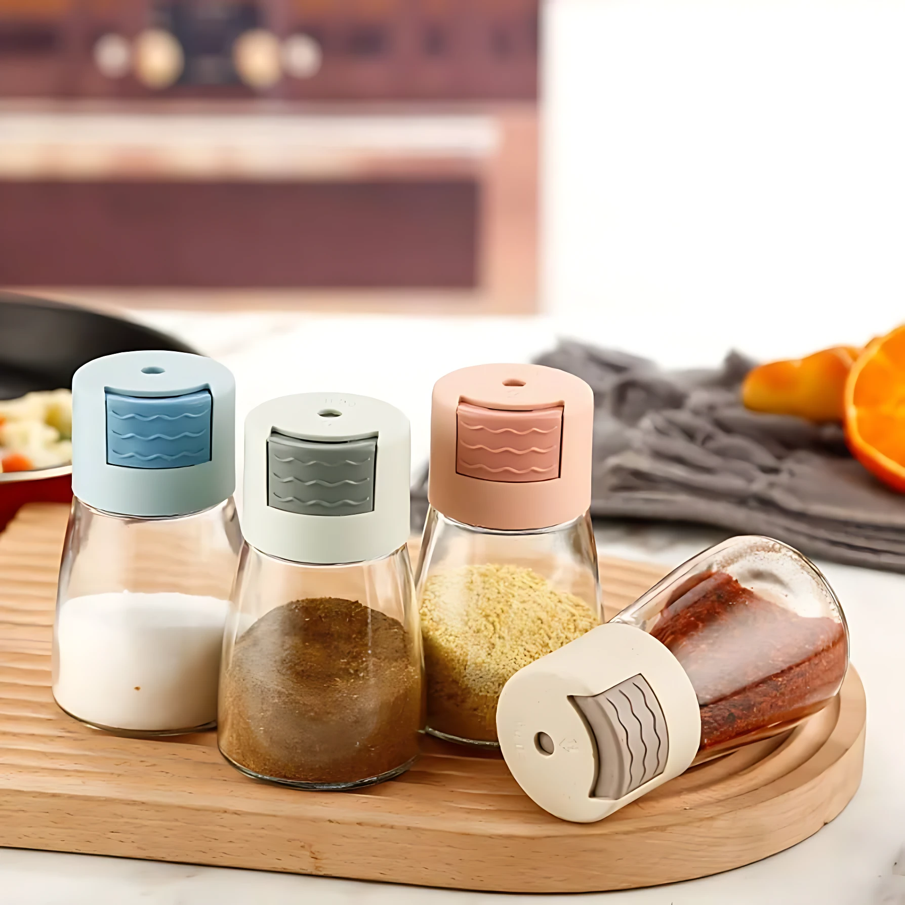Set de tres saleros de vidrio con tapa de plástico en azul, gris y rosa, cada uno con medidor de gramos integrado, dispuestos sobre tabla de madera junto a uno inclinado.
