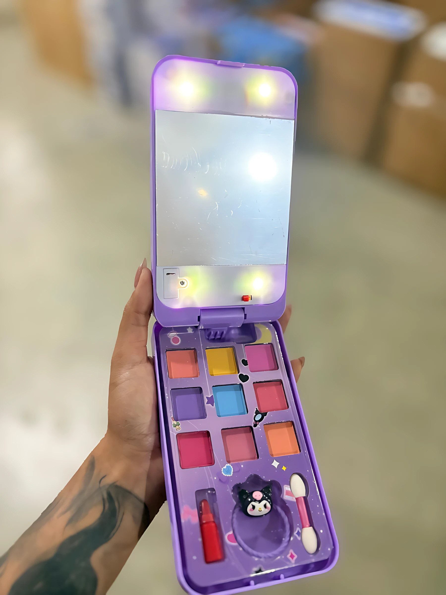 Set de maquillaje infantil morado con estuche tipo teléfono Kuromi, espejo iluminado, sombras multicolores, aplicador, labial y figura decorativa.
