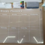 Tablero acrílico transparente con marco de cartón, incluye marcadores y adhesivos. Organizador semanal para nevera con días de la semana visibles.