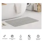 Tapete de baño gris con diseño ondulado, colocado en piso blanco junto a bañera y lavadora. Material de diatomita, textura suave y antideslizante.