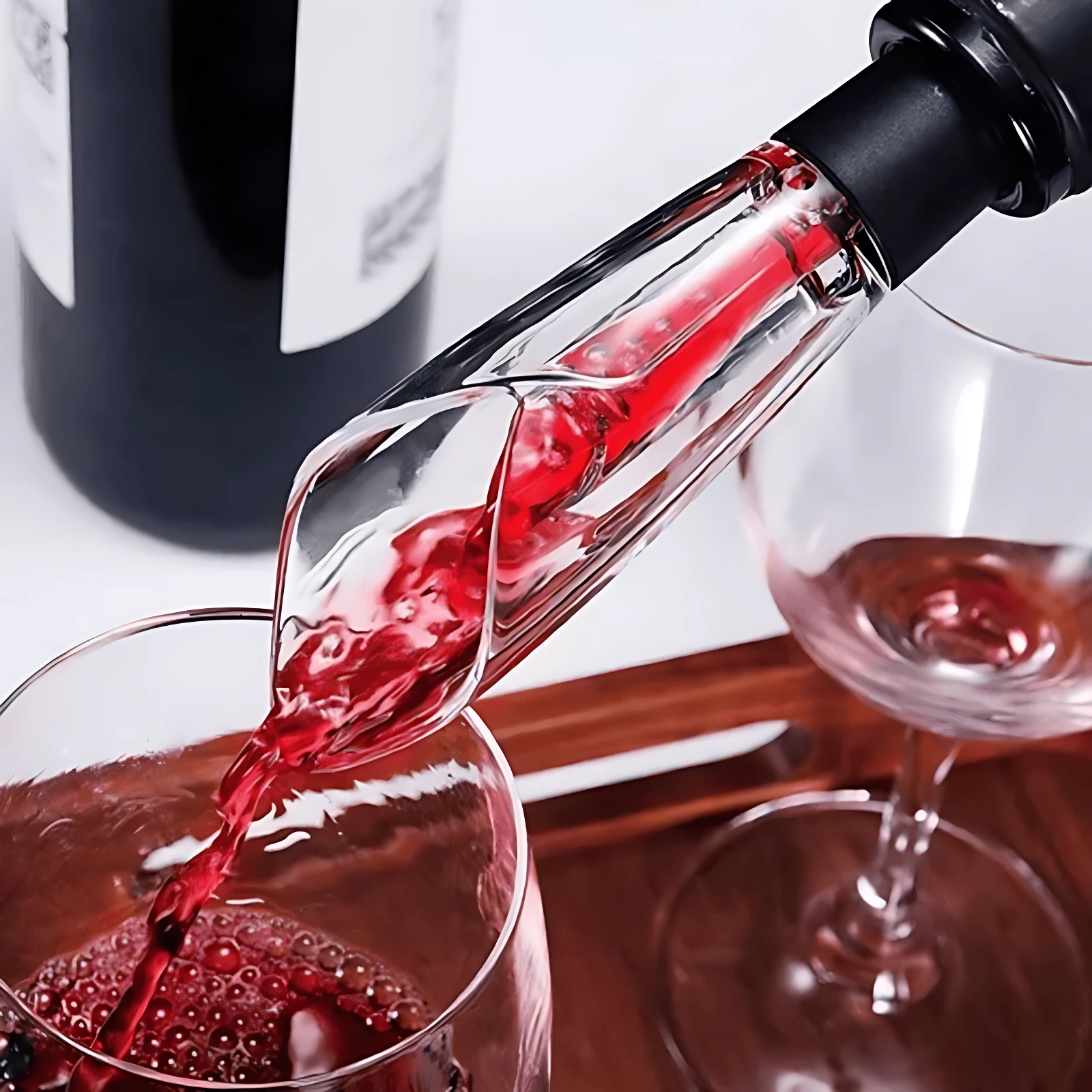 Tapón de vino filtrante en acción, vierte líquido rojo en copa con gotas visibles; fondo con botella y segunda copa vacía sobre bandeja de madera.