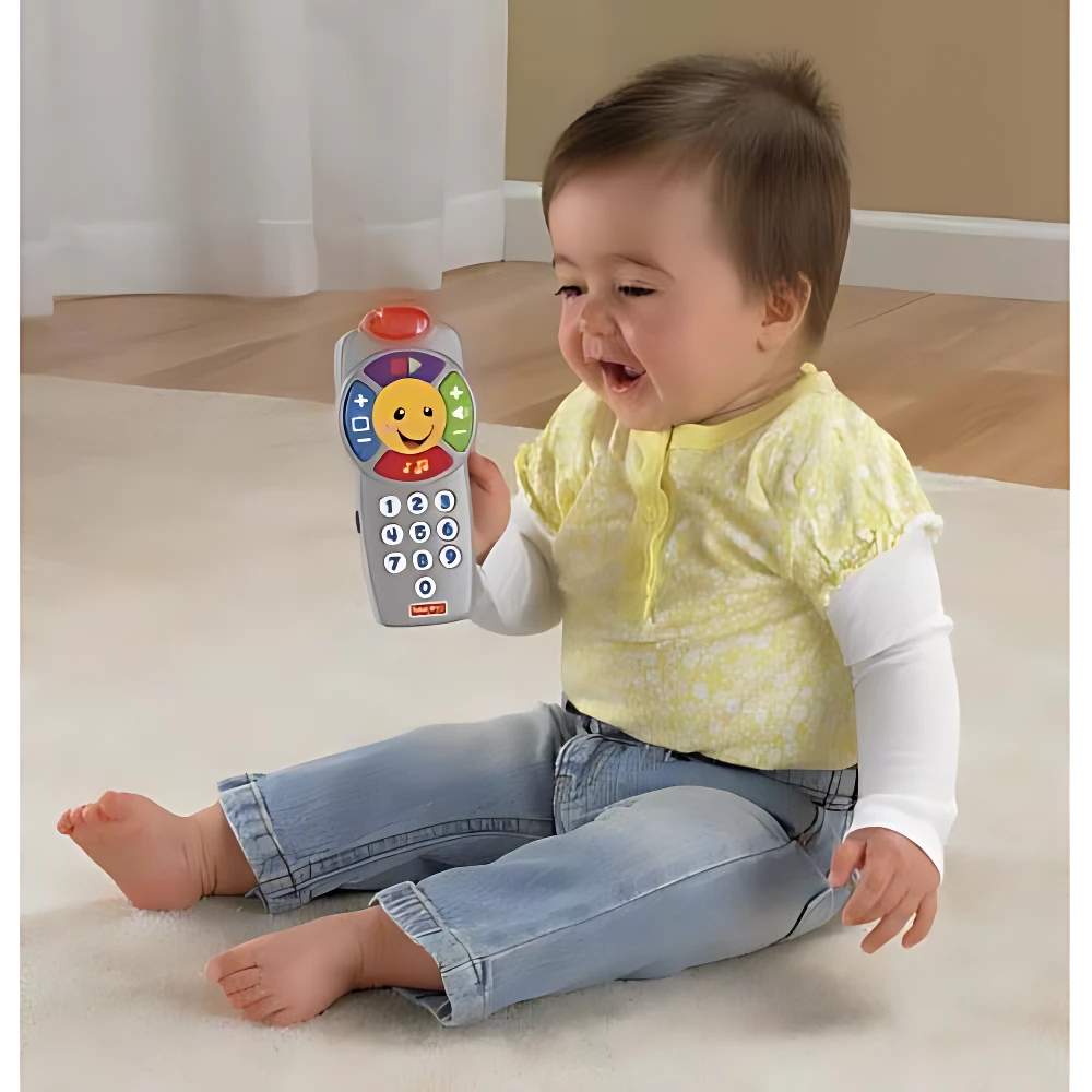 Bebé sentado en alfombra sosteniendo teléfono educativo interactivo gris con botones coloridos y pantalla solar, usado para estimular desarrollo cognitivo y motricidad fina.