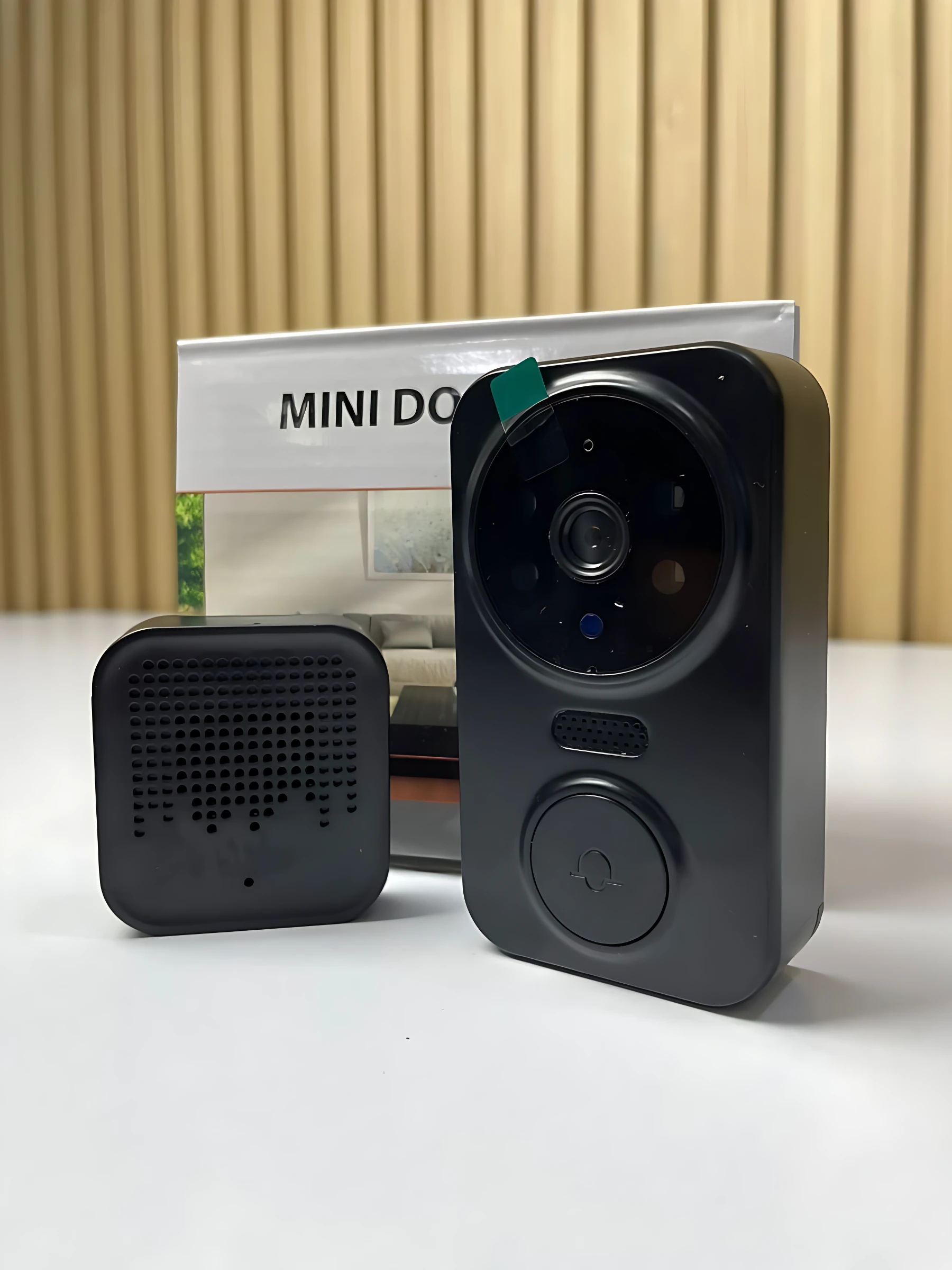 Kit de videoportero inalámbrico negro con unidad de cámara y timbre cuadrado con rejilla, junto a su caja blanca sobre fondo de madera.