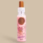 Perfume capilar rosa en frasco transparente con tapa bronce, de hada y sello decorativo sobre fondo beige.