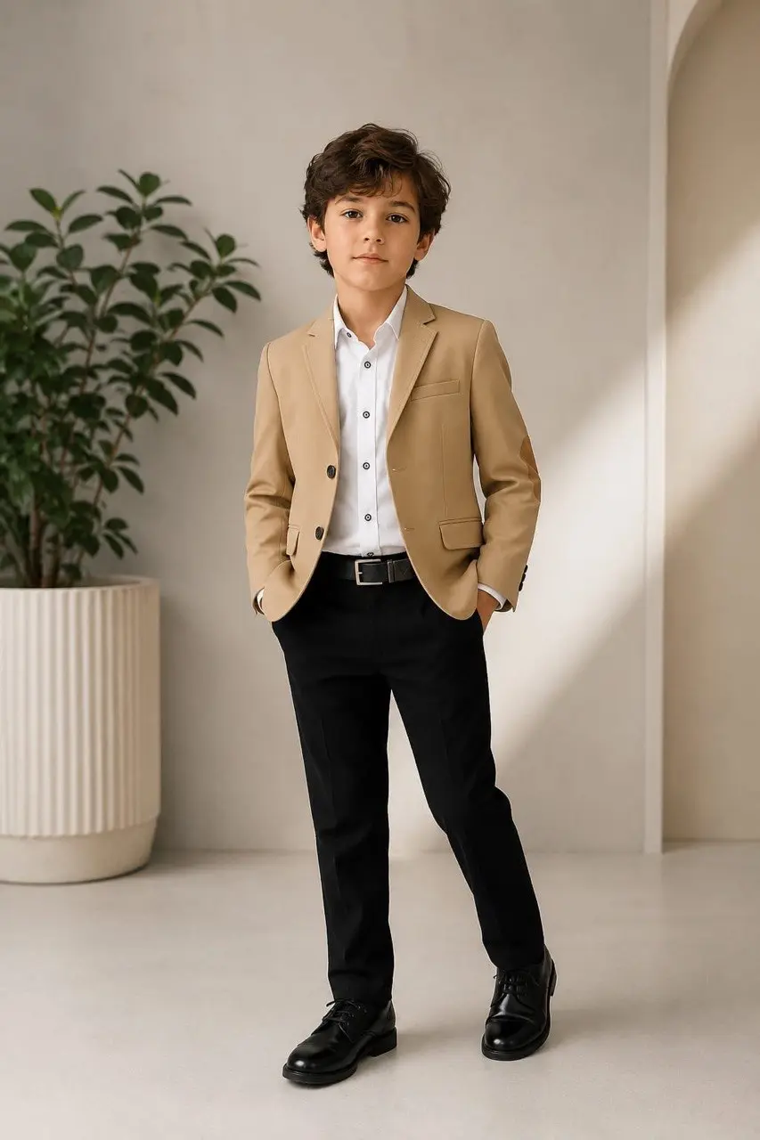 Niño de pie luciendo blazer caqui con coderas, camisa blanca, pantalón negro y zapatos formales en espacio interior con planta decorativa.