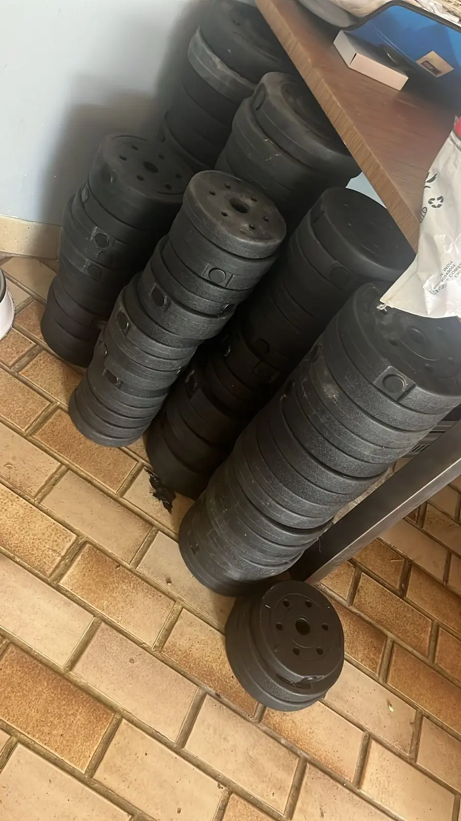 Discos de pesas negros de 3 kg apilados en columnas sobre un piso de baldosas, con un disco en primer plano junto a un escritorio de madera.