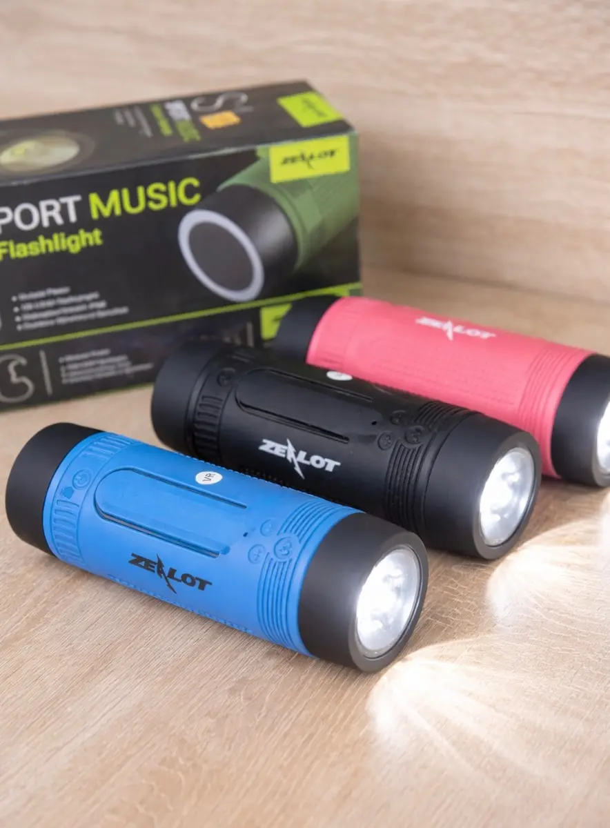 Parlantes Bluetooth Zealot S1 con linterna LED en colores azul, negro y rojo, con textura rugosa y luz encendida sobre superficie de madera.