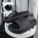 Par de sandalias deslizantes negras de suela EVA gruesa y texturizada, diseño ergonómico con ranuras en el empeine, exhibidas sobre una base blanca junto a una figura de astronauta.