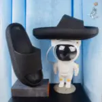 Pantuflas negras de suela EVA antideslizante con textura estriada, mostradas una vertical sobre base y otra horizontal sobre una lámpara de astronauta.